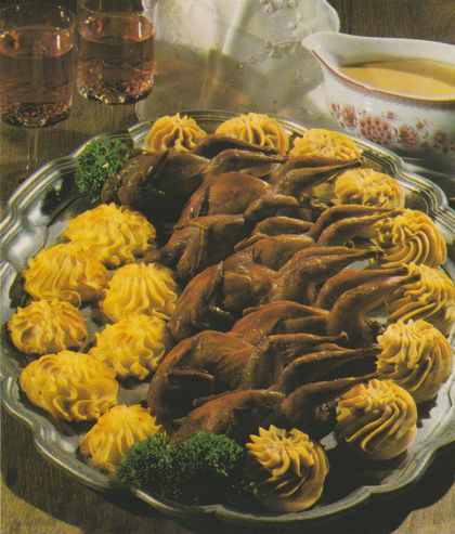 Festliche Servierplatte mit gebratenen Wachteln, umgeben von Artischockenböden und Herzogin-Kartoffeln, die mit goldgelben Kartoffelpüree-Rosetten dekoriert sind. Die Wachteln sind in der Mitte auf einer ovalen Glasplatte arrangiert, die gespritzten Kartoffel- und Maronenpüree-Tupfen bilden einen Kranz drumherum. Einzelne Petersilienzweige dienen als grüne Garnitur. Im Hintergrund sind Weingläser und eine Sauciere zu erkennen.