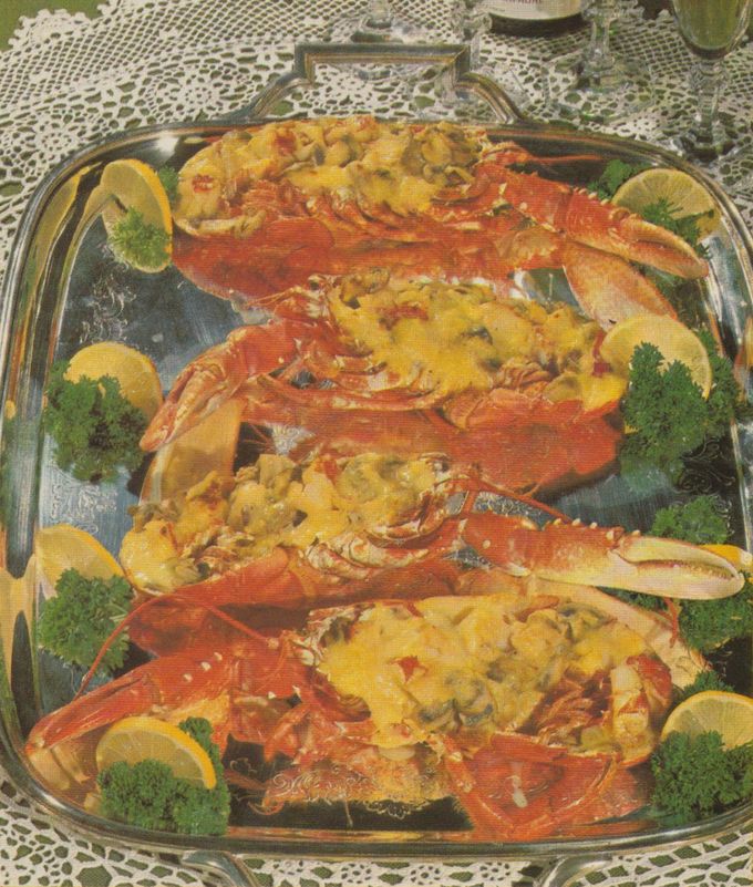 Vier halbierte Hummer auf einer ovalen Servierplatte, gefüllt mit einer Mischung aus Hummerfleisch, Champignons und Möhrenscheiben, überbacken mit einer Curry– oder Safransauce. Die Platte ist dekoriert mit Zitronenscheiben und Petersilie. Im Hintergrund eine Spitzendecke.