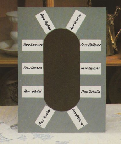 Eine selbstgebastelte Tischordnung aus grauem Karton mit einer ovalen Aussparung in der Mitte, um die herum zehn weiße Namensschilder für die Gäste angeordnet sind. Im Hintergrund sind verschwommen Geschirr und eine goldene Dekofigur zu erkennen.