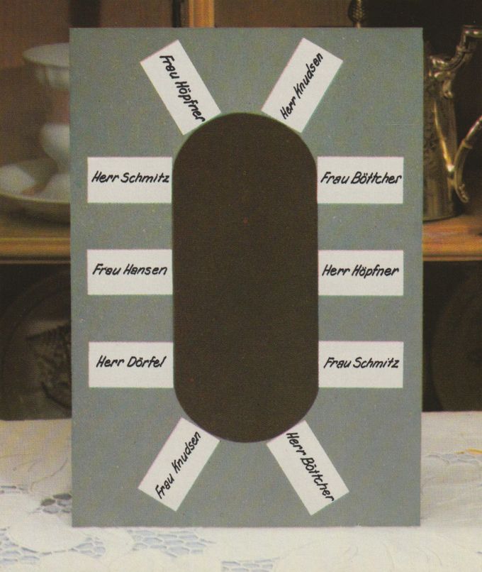 Eine selbstgebastelte Tischordnung aus grauem Karton mit einer ovalen Aussparung in der Mitte, um die herum zehn weiße Namensschilder für die Gäste angeordnet sind. Im Hintergrund sind verschwommen Geschirr und eine goldene Dekofigur zu erkennen.