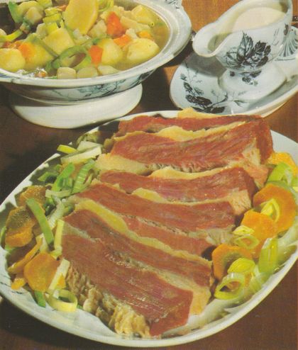 Aufgeschnittene gepökelte Rinderbrust auf einem Bett aus Lauch, Möhren und Kartoffeln auf ovalem Teller. Im Hintergrund eine Terrine mit Gemüse und eine Sauciere mit Meerrettichsauce, beide aus weißem Porzellan mit blauem Blumenmuster.