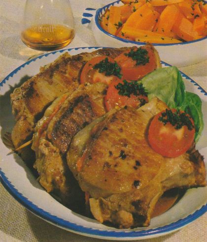 Vier gebratene, gefüllte Schweinekoteletts mit sichtbaren Grillstreifen auf einem weiß-blau geränderten Teller. Garniert mit Tomatenscheiben, gehackter Petersilie und einem Salatblatt. Im Hintergrund ein Glas Asbach Uralt sowie eine Schale mit gebutterten Möhren und Petersilie.