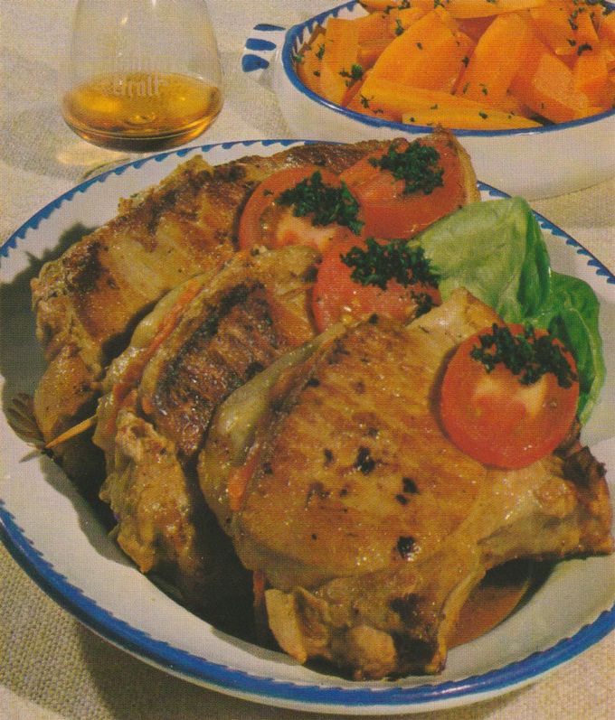 Vier gebratene, gefüllte Schweinekoteletts mit sichtbaren Grillstreifen auf einem weiß-blau geränderten Teller. Garniert mit Tomatenscheiben, gehackter Petersilie und einem Salatblatt. Im Hintergrund ein Glas Asbach Uralt sowie eine Schale mit gebutterten Möhren und Petersilie.