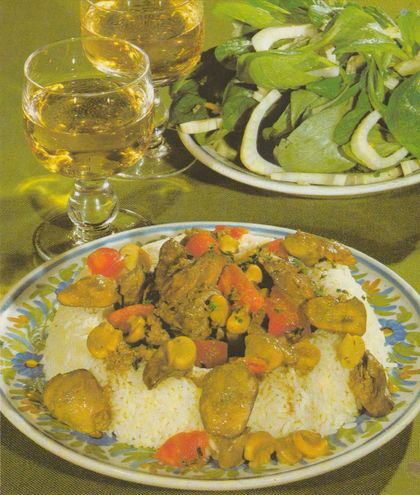 Teller mit Gericht aus Geflügelleber, Champignons und Tomatenstücken auf einem Reisbett, serviert auf einem Teller mit Blumenmuster. Im Hintergrund zwei Gläser Weißwein und eine Schüssel mit Feldsalat, garniert mit Zwiebelringen und Fenchel.