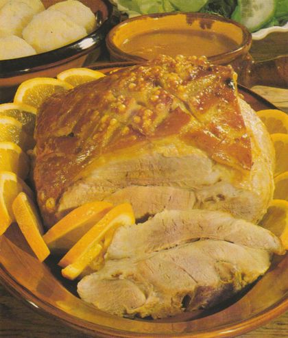 Aufgeschnittener Schweinebraten mit knuspriger, gitterförmig eingeschnittener Schwarte auf einem braunen Servierteller, umgeben von Orangen­scheiben und ‑spalten. Im Hintergrund eine Schüssel mit Klößen sowie ein Tonschälchen mit Orangen­sauce.