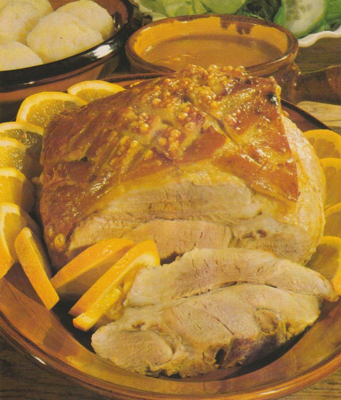 Aufgeschnittener Schweinebraten mit knuspriger, gitterförmig eingeschnittener Schwarte auf einem braunen Servierteller, umgeben von Orangen­scheiben und ‑spalten. Im Hintergrund eine Schüssel mit Klößen sowie ein Tonschälchen mit Orangen­sauce.