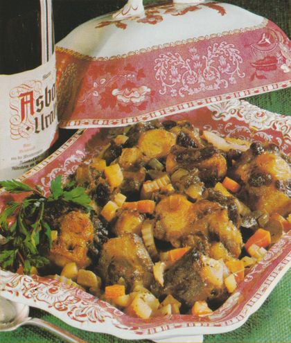 Geschmorter Ochsenschwanz mit Karotten, Sellerie und Zwiebeln in einer rot-weiß gemusterten Porzellanschale mit floralen Motiven. Links ein Krug Rotwein, daneben frische Petersilie als Garnitur. Im Hintergrund eine Falsche Asbach Uralt.