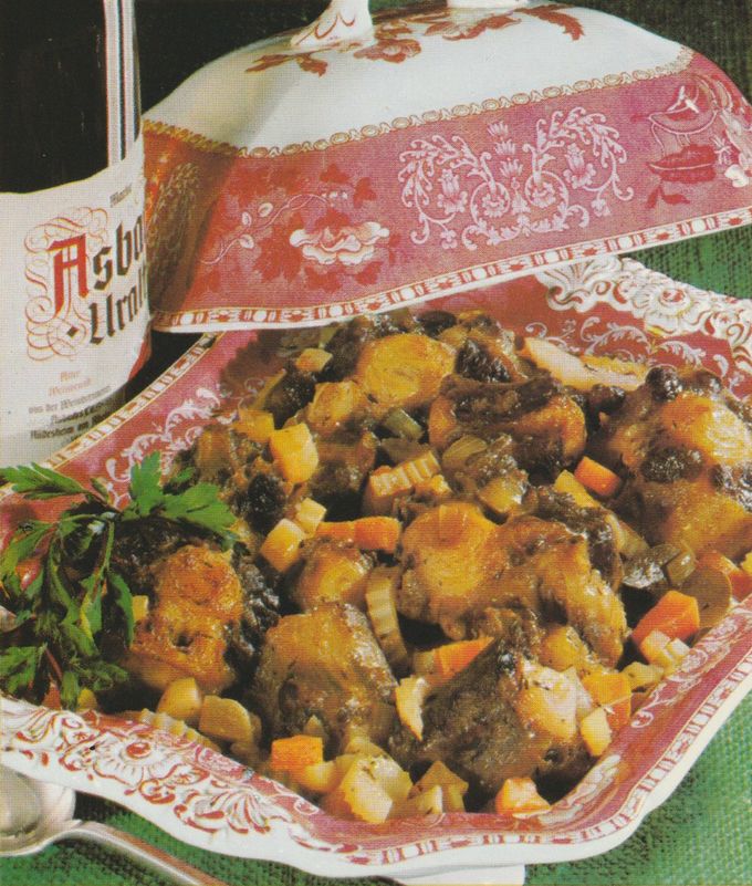 Geschmorter Ochsenschwanz mit Karotten, Sellerie und Zwiebeln in einer rot-weiß gemusterten Porzellanschale mit floralen Motiven. Links ein Krug Rotwein, daneben frische Petersilie als Garnitur. Im Hintergrund eine Falsche Asbach Uralt.