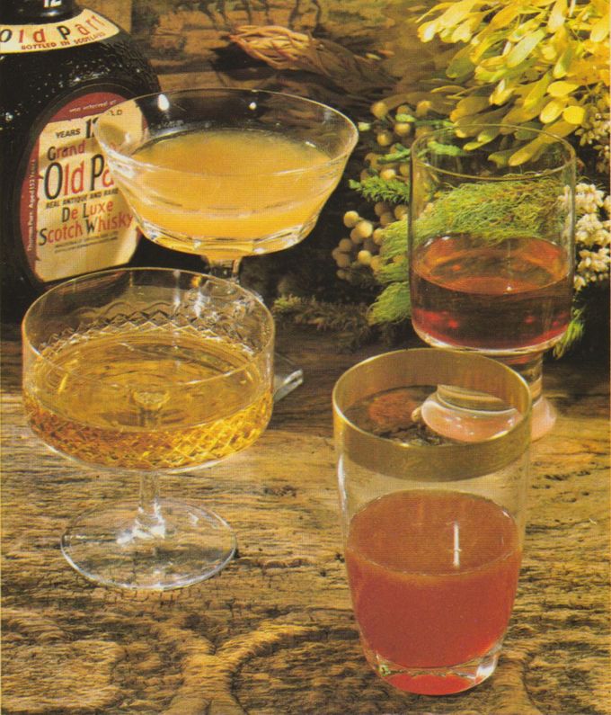 Vier verschiedene Whisky-Cocktails in unterschiedlichen Gläsern auf rustikalem Holztisch, im Hintergrund eine Flasche Scotch Whisky und gelbe Frühlingsblumen als Dekoration. Die Drinks reichen farblich von hellem Gold über Bernstein bis zu zartem Rosa.