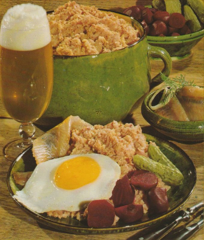 Portion Labskaus mit Spiegelei, Salzgurken, Rote Bete und Matjesfilet auf einem Teller. Im Hintergrund ein Bierglas, eine Schüssel mit weiterem Labskaus und eine Obstschale.