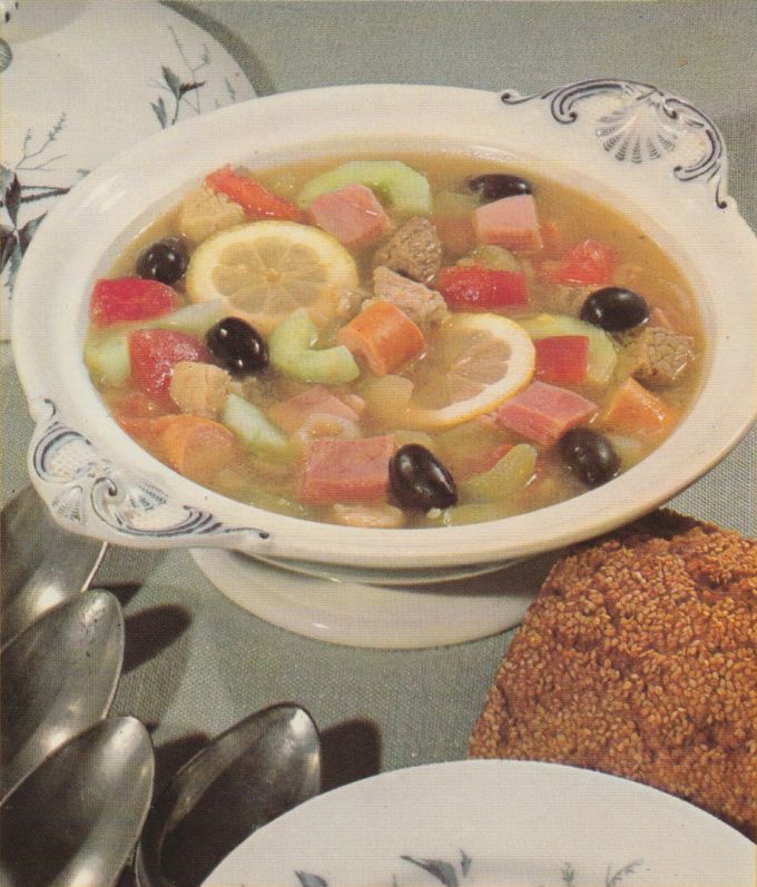 Eine klare Suppe mit Zitronenscheiben, Selleriestücken, Karotten, Fleisch und schwarzen Oliven wird in einer weißen Suppenterrine mit Schnörkeldekor serviert. Daneben liegen mehrere Löffel und ein mit Sesam bestreutes Brot.