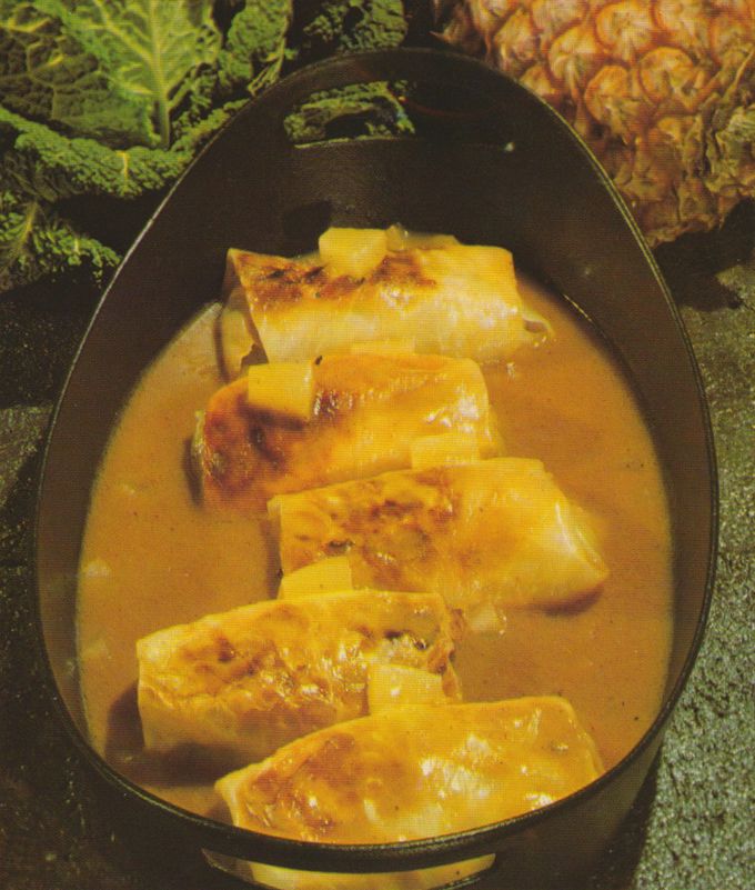 Gefüllte Kohlrouladen mit gebräunten Stellen liegen in einer cremigen, gelblichen Sauce in einem dunklen Topf. Im Hintergrund sind Wirsingkohlblätter und eine Ananas zu sehen.