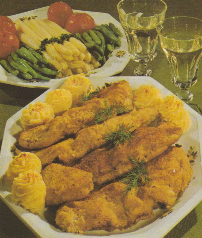 Panierte Kalbsschnitzel auf einer ovalen Servierplatte, garniert mit Dillzweigen und spritzgebackenen Kartoffelrosetten. Im Hintergrund eine Gemüseplatte mit Stangenspargel, grünen Bohnen, Maiskolben und Tomaten sowie Weißweingläser.