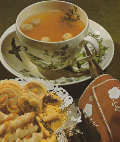 Eine Tasse klare Suppe mit Grießnocken schwimmt in einer vintage Porzellantasse mit Vogel- und Blumenmuster, dazu liegen Käsegebäck und Blätterteigschnecken auf einem Spitzendeckchen. Im Vordergrund sind braune Servietten mit weißer Blumenstickerei und Silberbesteck arrangiert.