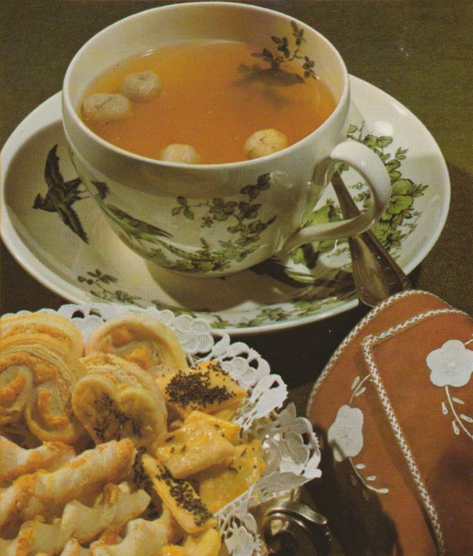 Eine Tasse klare Suppe mit Grießnocken schwimmt in einer vintage Porzellantasse mit Vogel- und Blumenmuster, dazu liegen Käsegebäck und Blätterteigschnecken auf einem Spitzendeckchen. Im Vordergrund sind braune Servietten mit weißer Blumenstickerei und Silberbesteck arrangiert.