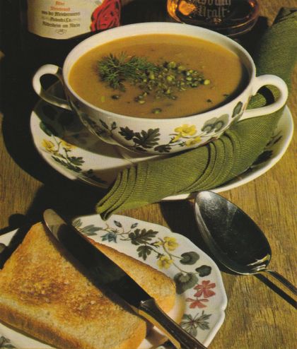 Eine cremige, goldgelbe Suppe in einer Suppentasse mit Blumenmuster, garniert mit frischen Kräutern und Maiskörnern, serviert auf grüner Stoffserviette mit geröstetem Brot und Löffel. Im Hintergrund sind weitere Geschirr-Elemente auf einem Holztisch zu erkennen.