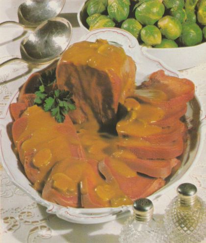 Aufgeschnittene gepökelte Rinderzunge in goldbrauner Madeira-Champignon-Sauce auf einem ovalen Teller mit rot-weißem Dekorrand, garniert mit Petersilie. Im Hintergrund eine Schüssel mit Rosenkohl, eine silberne Saucenkelle und Salz- und Pfefferstreuer auf weißer Tischdecke mit Spitzenmuster.